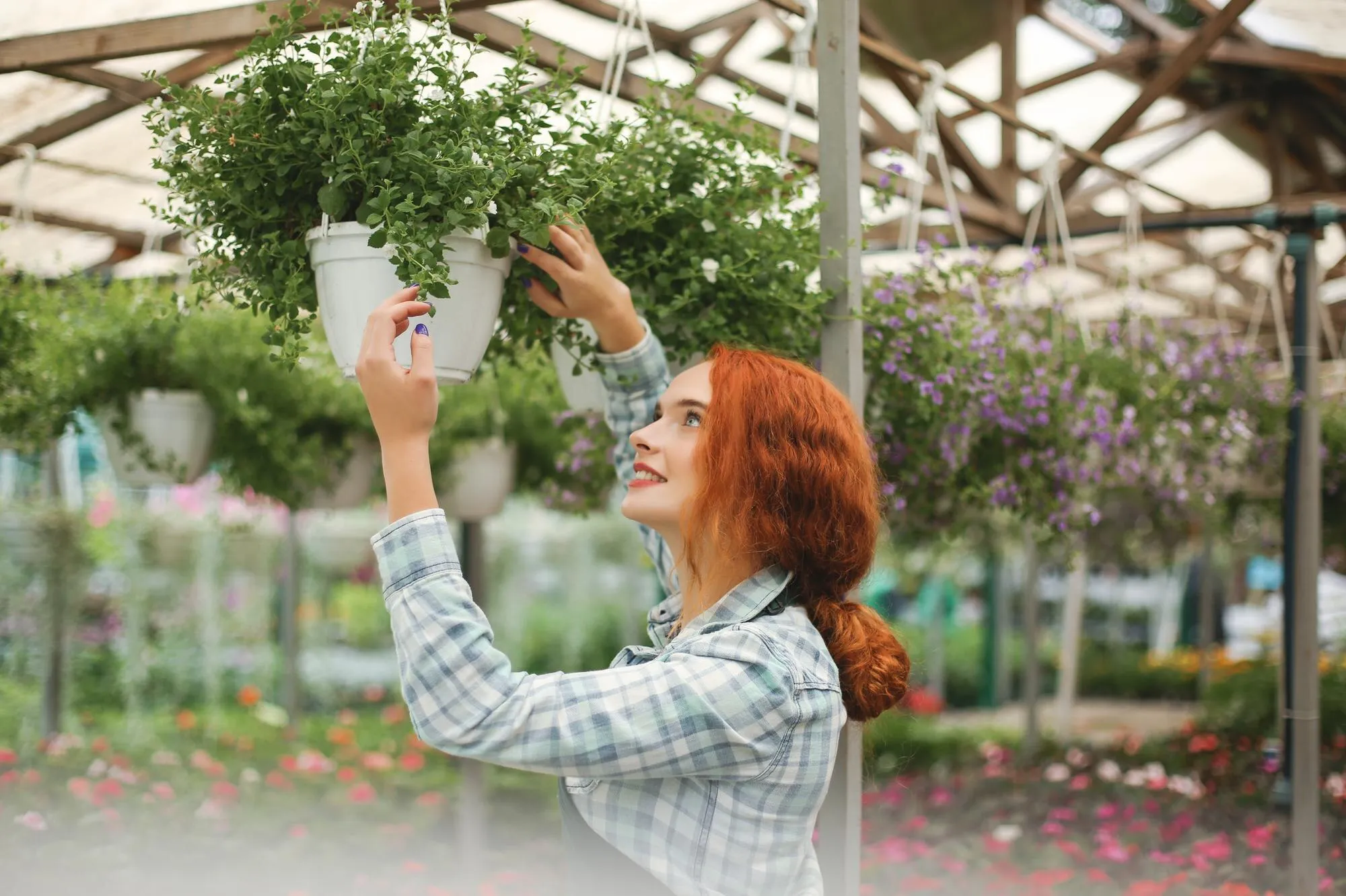 A white woman, 25 years old with red hair in a ponytail, wearing a blue and white shirt, touching a plant in a greenhouse.