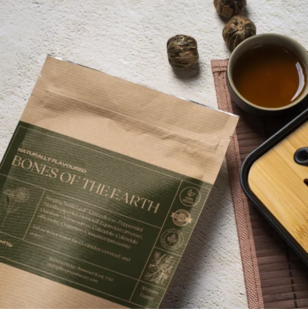 A flat-lay composition of a tea package labeled "Bones of the Earth" alongside a cup of brewed tea, some dried herbs or spices, and part of a bamboo or wooden tray.