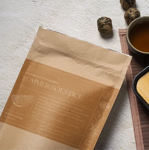 A bag of Summer Solstice tea in a brown paper kraft bag with a mustard yellow label, lying on a table, next to a cup of freshly brewed tea and a bamboo cup rest.