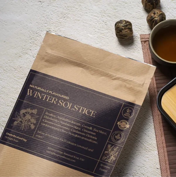 A bag of Winter Solstice tea in a brown paper kraft bag with a dark blue label, lying on a table, next to a cup of freshly brewed tea and a bamboo cup rest.