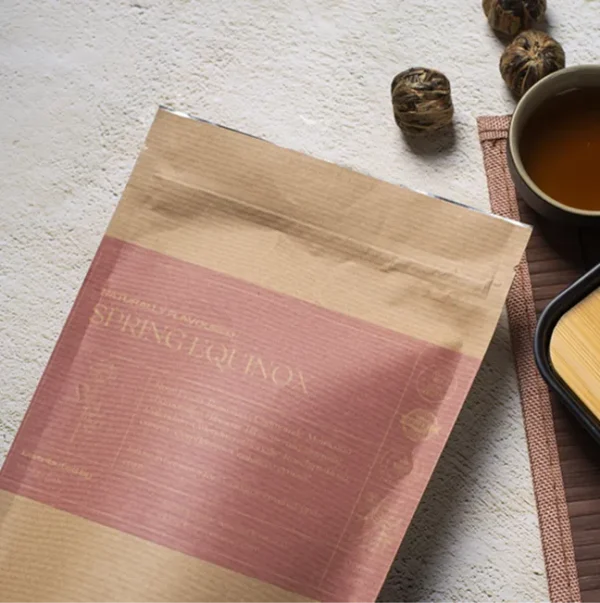 A bag of Spring Equinox tea in a brown paper kraft bag with a baby pink label, lying on a table, next to a cup of freshly brewed tea and a bamboo cup rest.