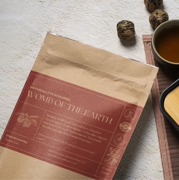 A bag of Womb of the Earth tea in a brown paper kraft bag with a red label, lying on a table, next to a cup of freshly brewed tea and a bamboo cup rest.