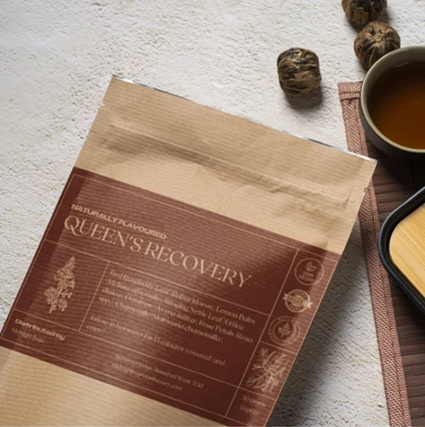 A bag of Queen's Recovery tea in a brown paper kraft bag with a dark red label, lying on a table, next to a cup of freshly brewed tea and a bamboo cup rest.