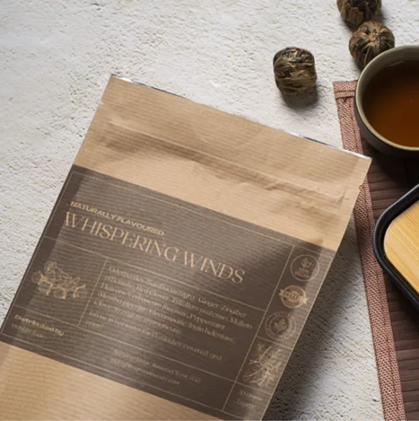 A bag of Whispering Winds tea in a brown paper kraft bag with a grey label, lying on a table, next to a cup of freshly brewed tea and a bamboo cup rest.