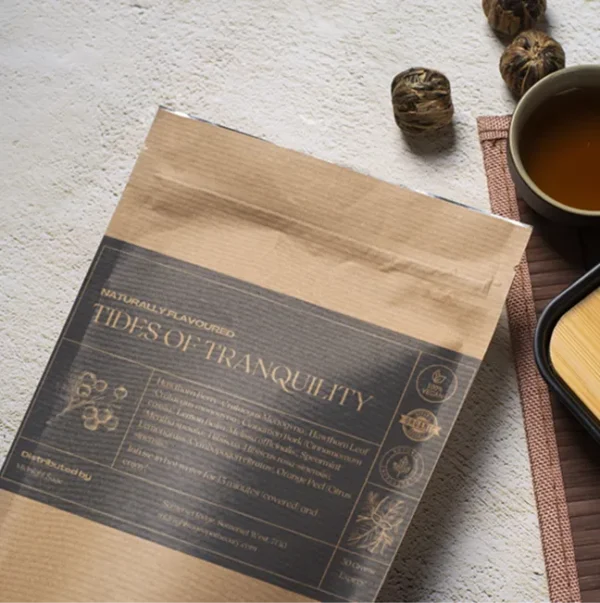 A bag of Tides of Tranquility tea in a brown paper kraft bag with a dark grey label, lying on a table, next to a cup of freshly brewed tea and a bamboo cup rest.