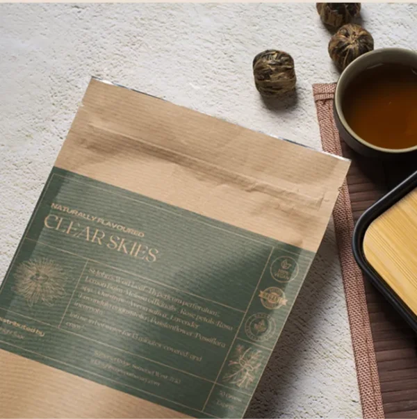 A bag of Clear Skies tea in a brown paper kraft bag lying on a table, next to a cup of freshly brewed tea and a bamboo cup rest.