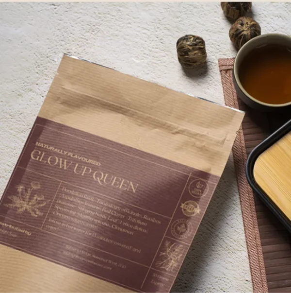 A bag of Glow Up Queen tea in a brown paper kraft bag with a purple label, lying on a table, next to a cup of freshly brewed tea and a bamboo cup rest.