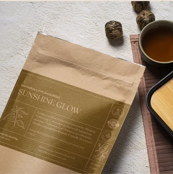 A bag of Glow Up Queen tea in a brown paper kraft bag with a yellow label, lying on a table, next to a cup of freshly brewed tea and a bamboo cup rest.