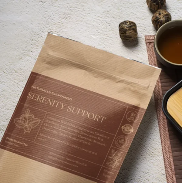 A bag of Serenity Support tea in a brown paper kraft bag with a maroon label, lying on a table, next to a cup of freshly brewed tea and a bamboo cup rest.