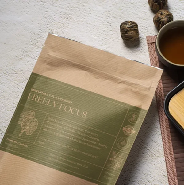 A bag of Freely Focus tea in a brown paper kraft bag with a dark light green label, lying on a table, next to a cup of freshly brewed tea and a bamboo cup rest.