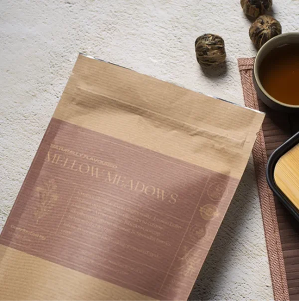 A bag of Mellow Meadows tea in a brown paper kraft bag with a light purple label, lying on a table, next to a cup of freshly brewed tea and a bamboo cup rest.