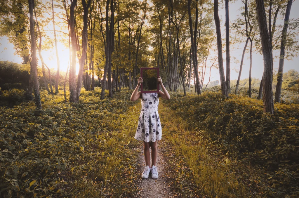Girl wearing a blue and white dress walking through a forest with the sun shining through the branches